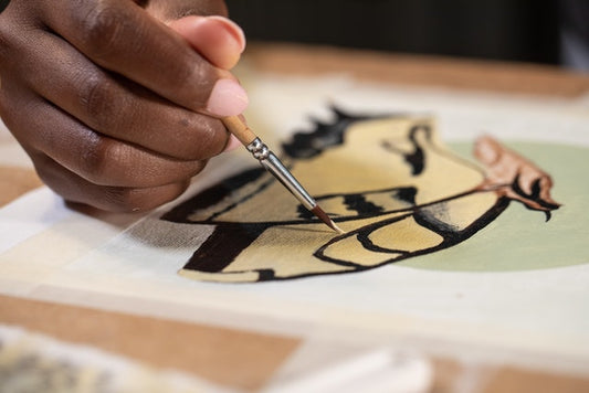 Close-up of a hand using a fine paintbrush to add details to a painting. The artwork features abstract shapes and lines in shades of black, beige, and green.