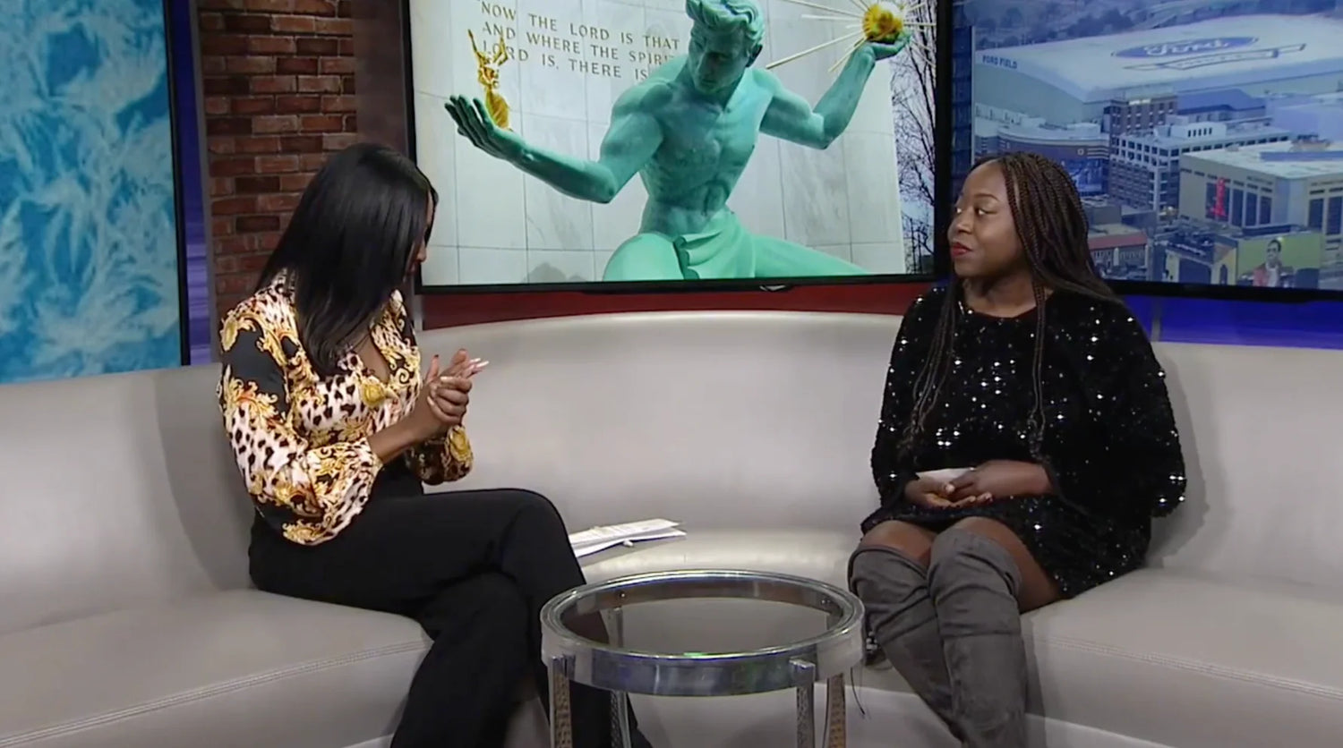 Two women sitting on a gray couch in a TV studio. One is talking with hand gestures, wearing a patterned top; the other is listening, wearing a black dress. A sculpture and cityscape are visible on a screen in the background.