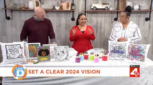 Three people stand at a table displaying coloring books, completed artworks, and markers. A banner reads SET A CLEAR 2024 VISION. The setting appears to be a TV studio with craft items and decor in the background.