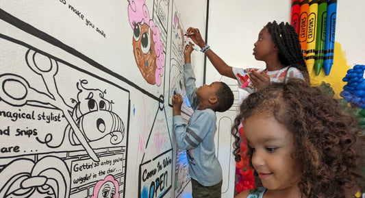 Children coloring on a large, black and white illustrated wall. A girl is reaching up with a marker, a boy is coloring closer to the floor, and another girl is smiling in the foreground. Large crayon decorations are on the side.