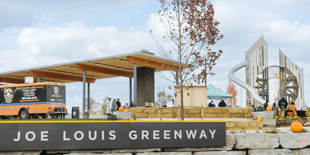 A scenic view of the Joe Louis Greenway featuring a modern pavilion, a food truck, and a decorative installation. People are present, and the area is adorned with pumpkins, hay bales, and trees.