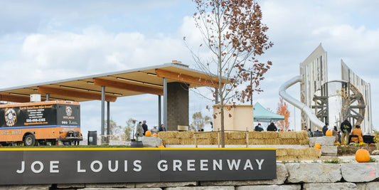 A scenic view of the Joe Louis Greenway featuring a modern pavilion, a food truck, and a decorative installation. People are present, and the area is adorned with pumpkins, hay bales, and trees.