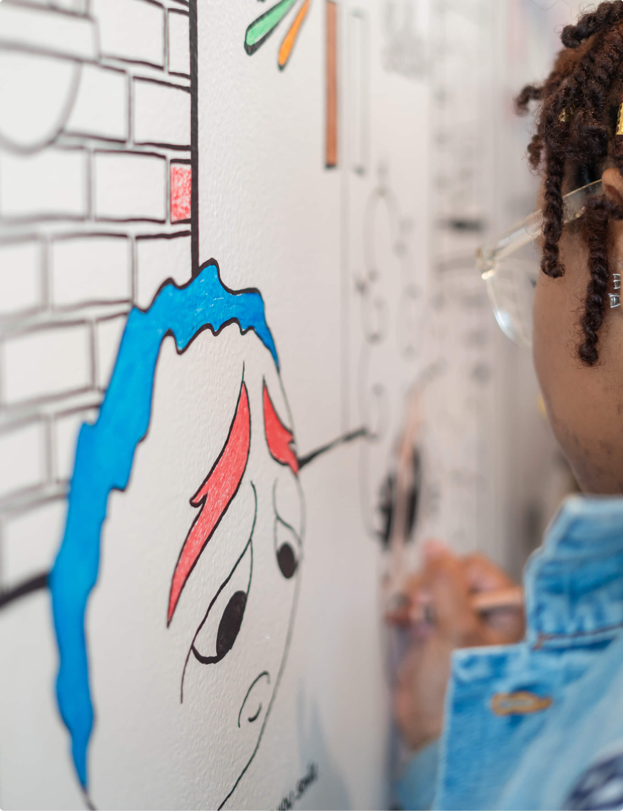 A person with glasses and braided hair draws on a white wall, adding color to a cartoon face with red eyebrows. The wall features various sketches, including a brick pattern. The person is wearing a denim jacket.