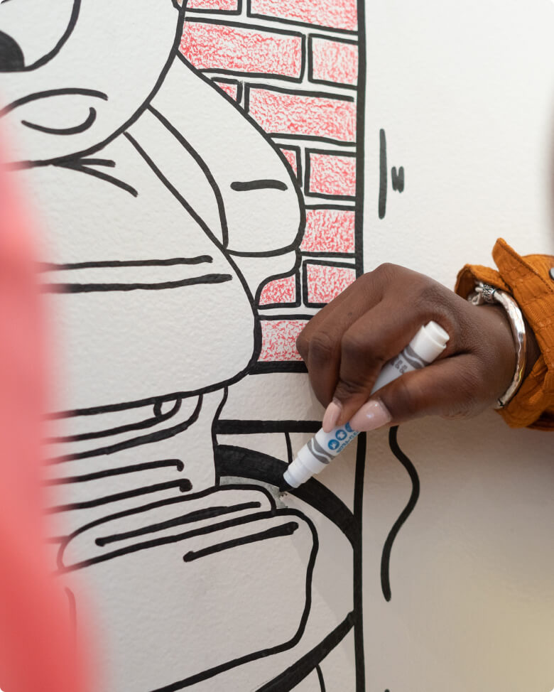 A person drawing a cartoon character with a black marker on a white wall. The character is outlined with bold lines, and a brick pattern is visible in the background. The persons hand is partially visible, holding the marker.