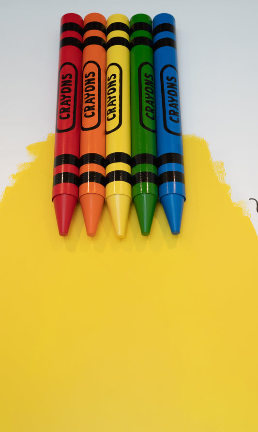 Five oversized crayons in red, orange, yellow, green, and blue are aligned horizontally on a yellow surface. Each crayon has the word CRAYONS printed on its side in black.